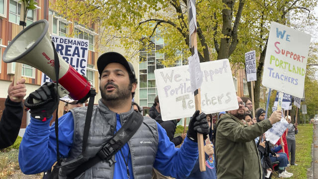 Teachers and their supporters are seen holding signs and rallying in support of a teacher's strike in Portland, Oregon. A man holding a sign and a bullhorn is in the foreground. 