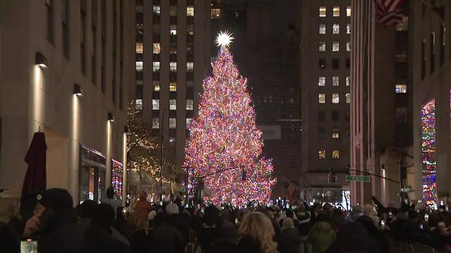 Dozens of people bundled up in winter gear walk behind barricades towards the Rockefeller Center Christmas tree. 