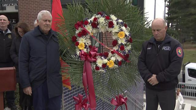 Two officials stand on either side of a flowered wreath, closing their eyes during a moment of silence. 