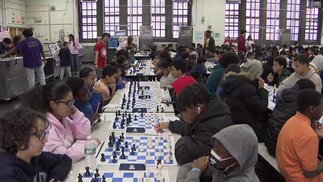 Dozens of children sit at chess tables. 