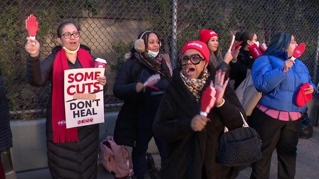 Half a dozen protesters stand outside an Inwood hospital. One holds a sign reading, "Some cuts don't heal." 