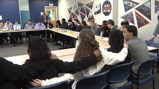 Students sit in a circle at long tables with their arms around each other's shoulders. 