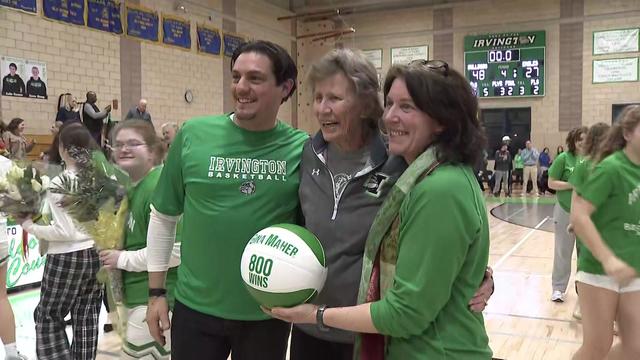 Coach Gina Maher poses for a photo between a man and a woman. The woman holds a basketball that says "Gina Maher 800 wins." 