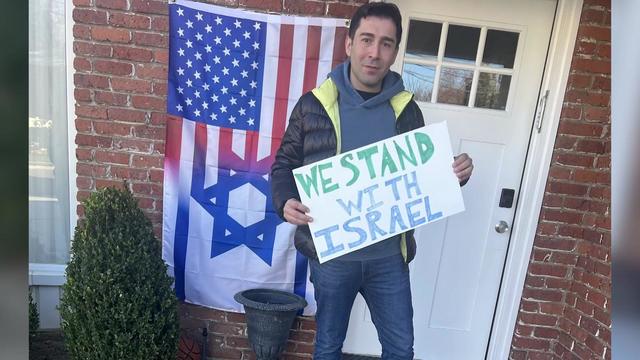 Aleks Binyaminov stands in front of his home holding a sign that says, "We stand with Israel." A combination American-Israeli flag hangs next to the front door behind him. 