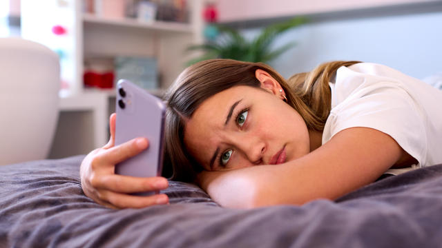 Depressed Teenage Girl Lying On Bed At Home Looking At Mobile Phone 