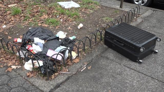 A bag of trash lays in a patch of grass outside a New York City park and a suitcase lays on the sidewalk. 