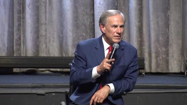 Texas Gov. Greg Abbott holds a microphone and sits in a wheelchair in front of a stage. 