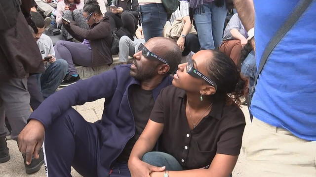 Two people in a crowd are wearing eclipse glasses and looking up at the sky 