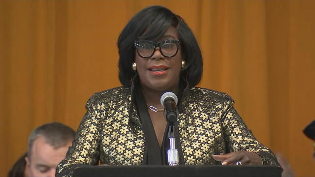 Mayor Cherelle Parker speaks into a microphone at a podium, she's wearing a black and gold jacket 