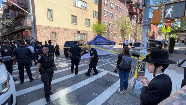 A black SUV sits on a street near a white sheet on the ground and a tent. The street is blocked off by crime scene tape and police officers push people back. 
