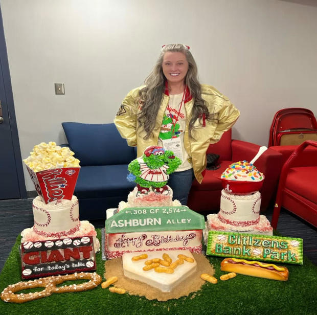 The baker stands with her cake, which is made up of ballpark foods including popcorn, a soft pretzel, ice cream in a small batting helmet and a hot dog 