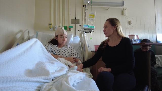 Maxi Park (left) sits in a hospital bed with a bandage around her head. She holds hands with her mother, who is sitting in a chair next to the bed. 