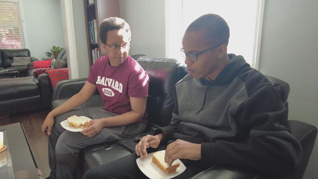 Emmanuel Gitu sits on the couch with his brother, Ian. They have peanut butter and jelly sandwiches on plates on their laps. 