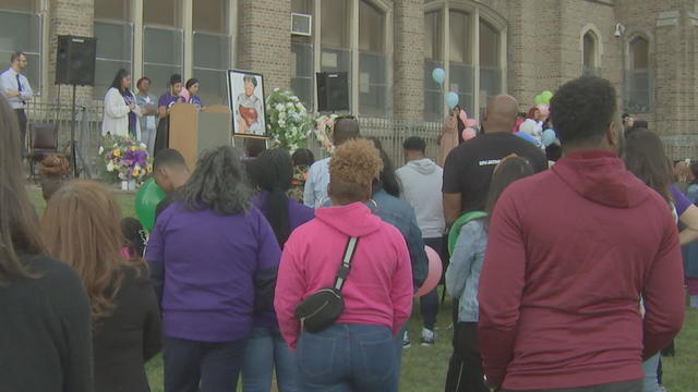 A crowd gathers for the vigil, people stand at a podium in the front, next to a portrait of Ondria Glaze and flowers 