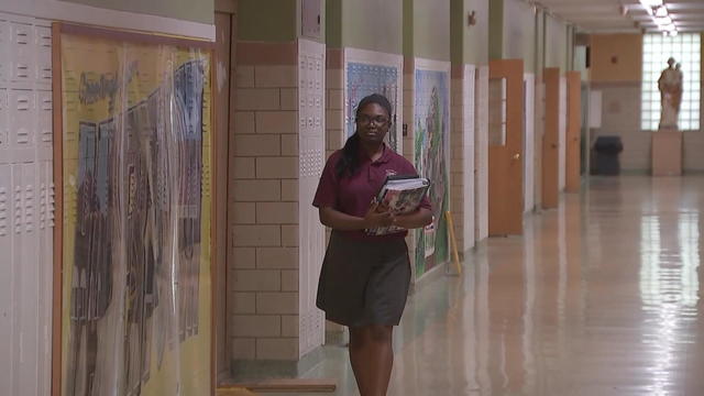A high school student holding books walks down a school hallway 