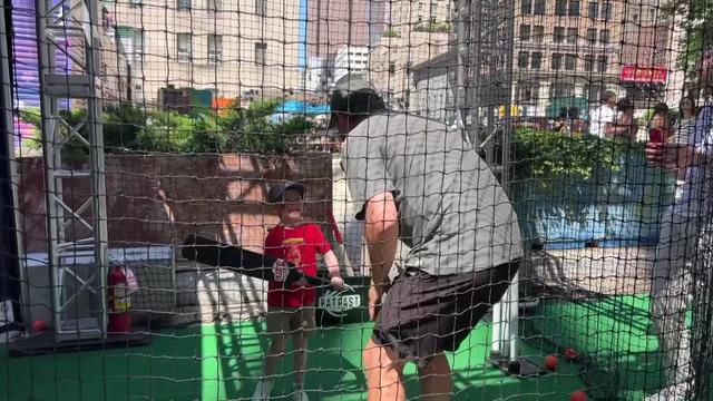 A child holds a cricket bat in a cricket simulator batting cage and smiles at an adult. 