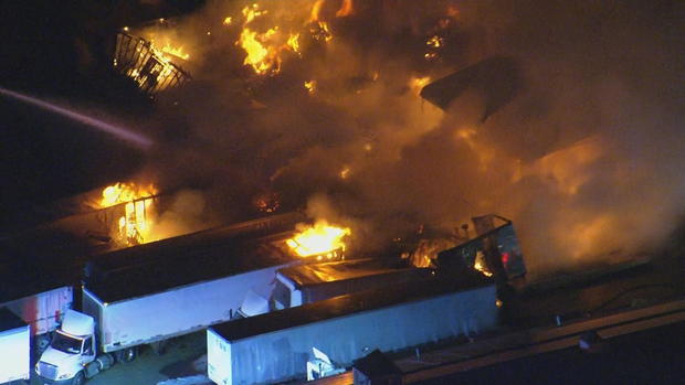 Flames are seen coming from trailers and pallets, a stream of water moves toward the fire 