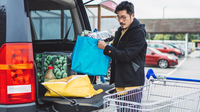 Young man putting away bags of groceries in his car trunk at supermarket car park 