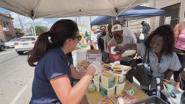 A volunteer holds out a plate holding two cheeseburgers while a member of the community adds ketchup 