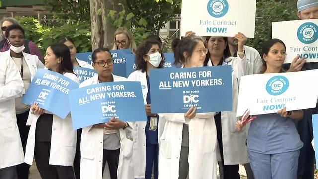 Health care workers holding signs reading "Caring for all New Yorkers" and "Fair contract now" stand outside a hospital. 