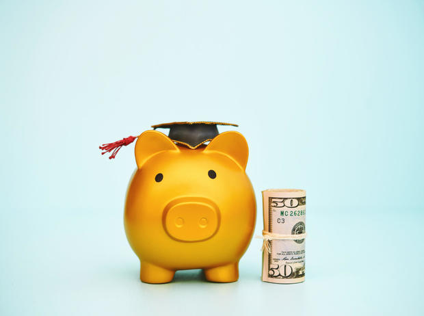 Cute gold colored piggy bank wearing a graduation cap and standing next to a roll of dollars. Education cost theme 