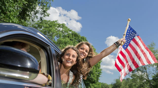 Two friends leaning out of car window holding american flag 