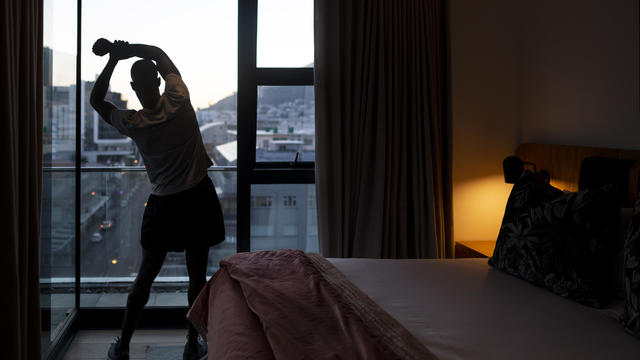Young man stretching by bedroom window looking at city view at sunrise 
