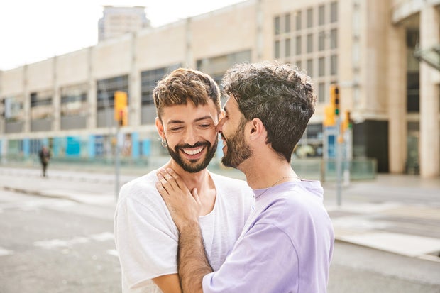 A gay couple share smiles and an intimate moment on a city street.