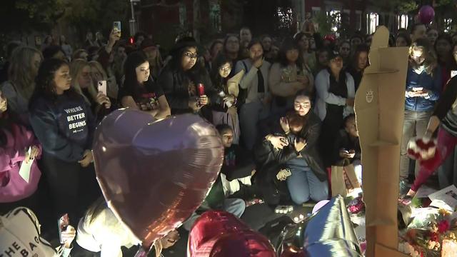 Hundreds of people stand and kneel in front of a makeshift memorial with flowers, balloons and a cardboard cutout of Liam Payne. 