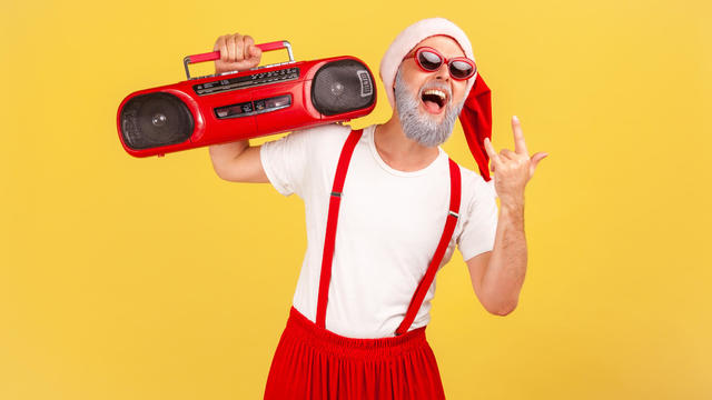 Funny happy gray bearded man in santa claus hat and stylish sunglasses holding tape recorder showing rock and roll gesture, listen to music 