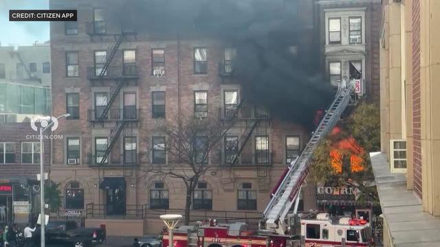 Flames and black smoke billow from a second-floor window of an apartment building. 