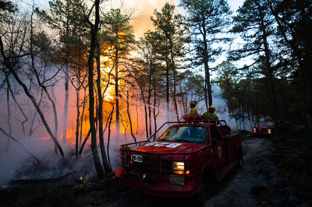 Firefighters are seen in a truck spraying water toward flames in a forest 