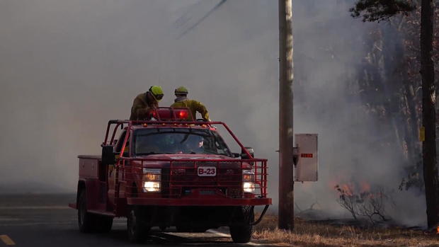 Firefighters are on a firetruck as smoke billows in the background 