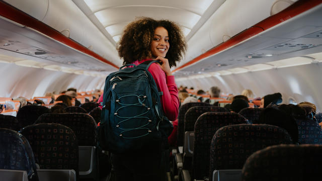 A woman boards a commercial aircraft with hand luggage. She glances over her shoulder and smiles at the camera. 