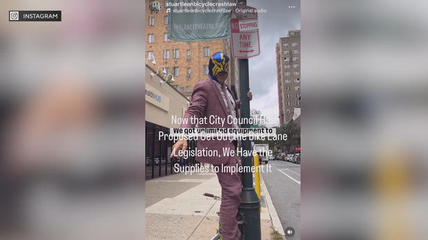 A man wearing a luchador mask hangs from a signpost after putting a sticker to change the sign from No Parking to No Stopping 