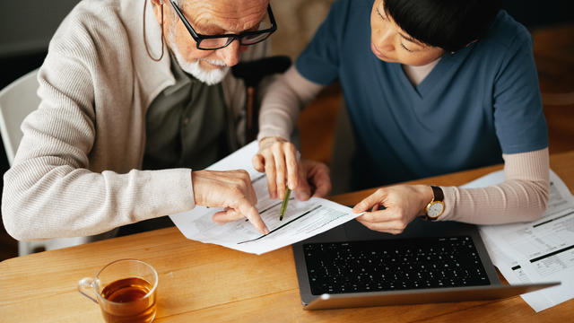 From Above Photo Of A Senior Man Using A Laptop Computer And Paying Bills With Serious Nurse 