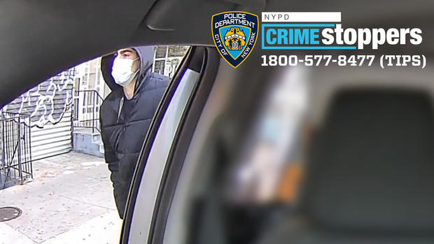 A man wearing a face mask and a black jacket with the hood up walks outside a vehicle. 