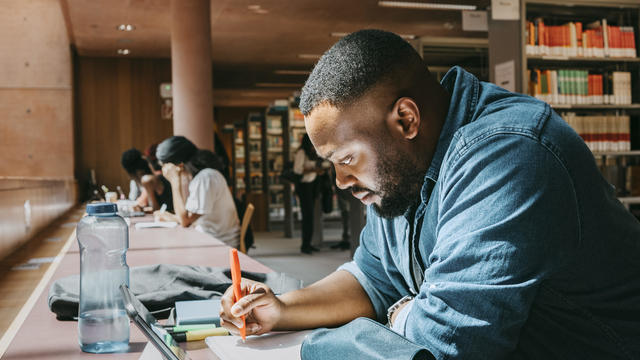 Student writing in notebook while sitting with tablet PC in college library 