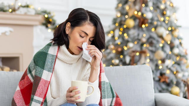 A young woman fell ill during the Christmas and New Year holidays. She sits at home on the sofa covered with a blanket, wipes her nose from a runny nose with a napkin, holds a cup of drink, medicine 