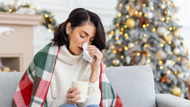 A young woman fell ill during the Christmas and New Year holidays. She sits at home on the sofa covered with a blanket, wipes her nose from a runny nose with a napkin, holds a cup of drink, medicine