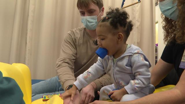 A medical student works with an 18-month-old child playing with a toy, along with a therapist. 