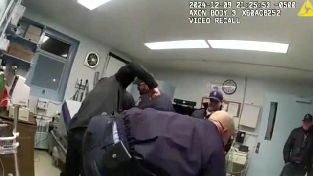 Correctional officers surround an inmate on a medical exam table. One pulls back his fist to punch him. 