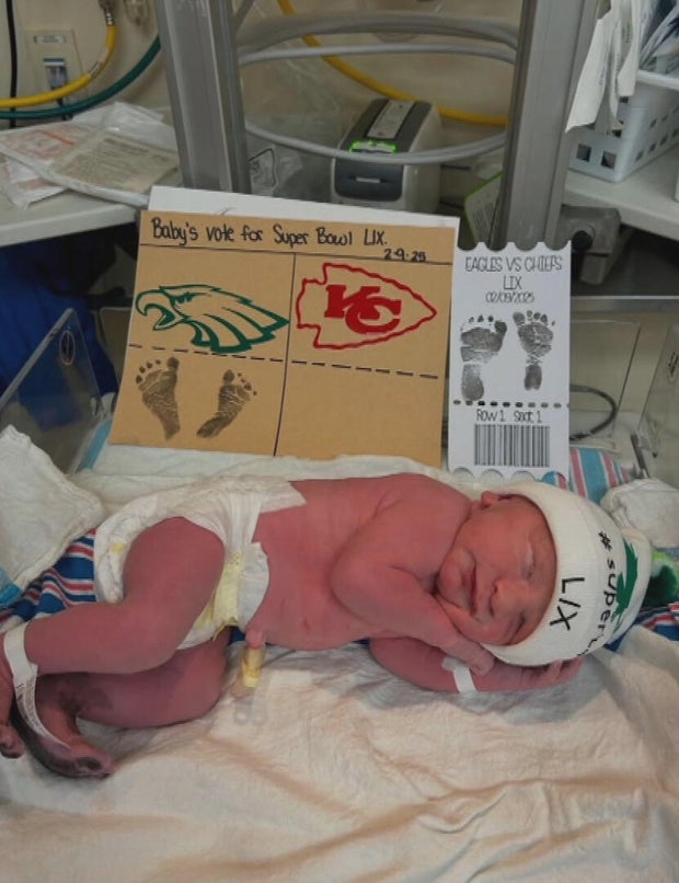 A baby wearing a hat that says Super Bowl LIX, a sign behind him says he is choosing the Eagles