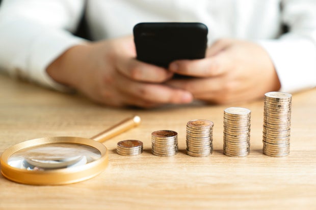 Businessman is opening data on his mobile phone to earn more money with magnifying glass and pile of money on the table. 