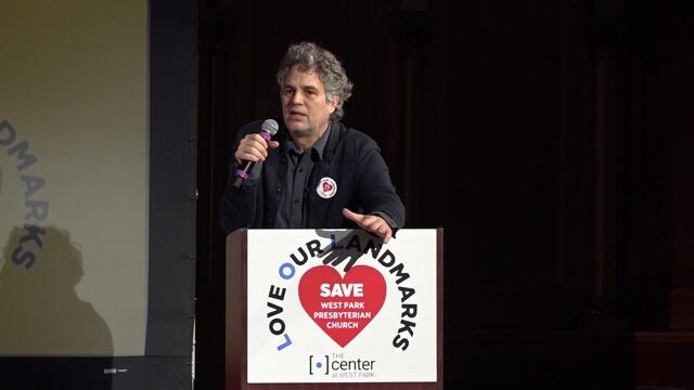 Mark Ruffalo stands a podium speaking into a microphone 