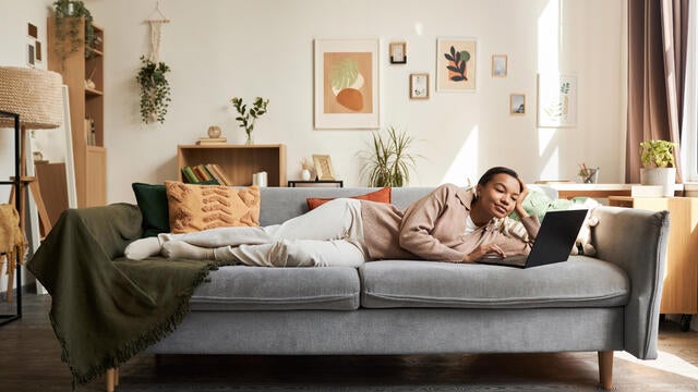 Young Black woman lying on couch in cozy home interior and using laptop 