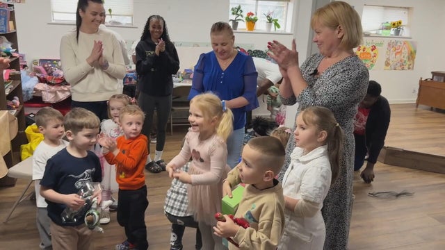 Young children clap and smile, adults stand nearby doing the same 