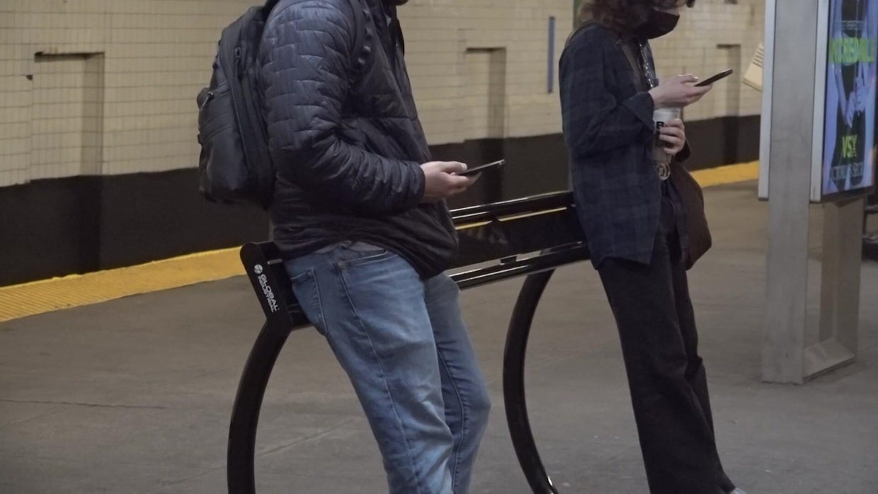 Two individuals lean against a slightly angled black metal "leaning bench" on a subway platform. 