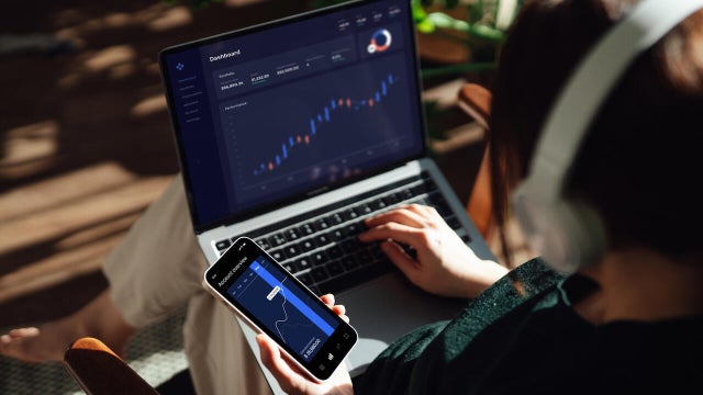 Over the shoulder view of young woman making financial plans and investment analysis via laptop and smartphone 