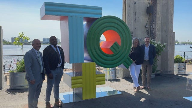 The sculpture is a T and a Q on top of a plus sign, in this photo, people post for a photo standing on either side of it 